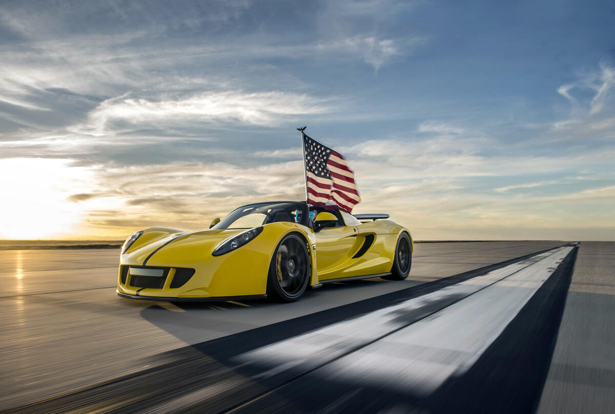 A yellow Hennessey Venom GT drives down the runway with an American flag flying in the wind