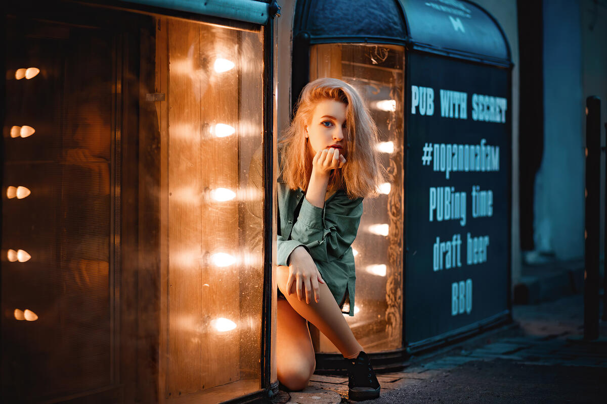 A young blonde looks out from behind a glowing corner of a store window
