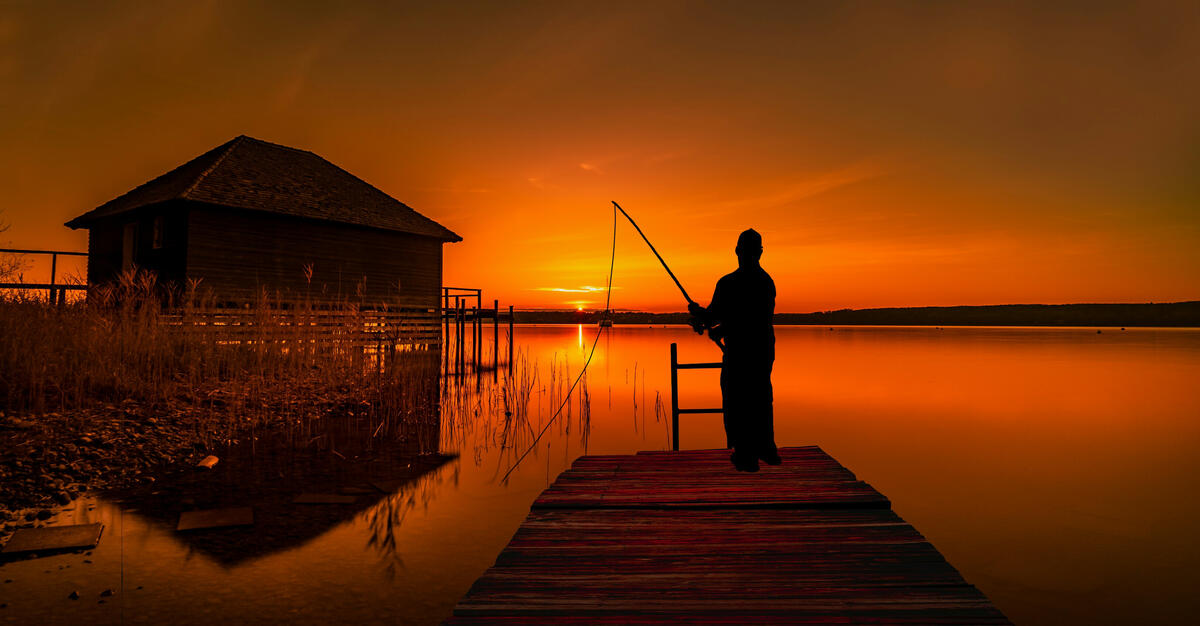 Силуэт рыбака на деревянном мостике рядом с деревянным домиком на закате