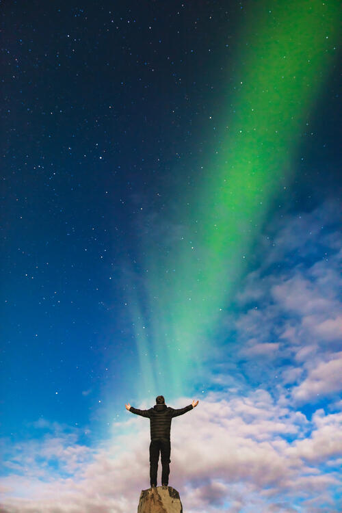 A man admires the northern lights while standing on the edge of a cliff.