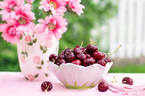 A plate of cherries on a table with a vase of pink flowers