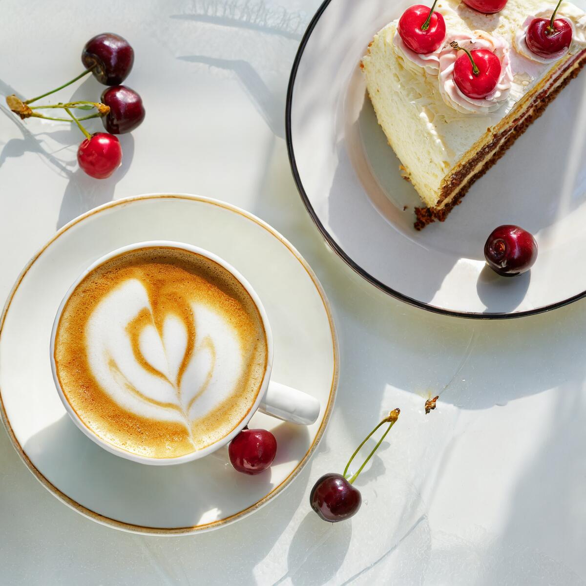 A white cup of cappuccino coffee next to a plate with a slice of cake on it