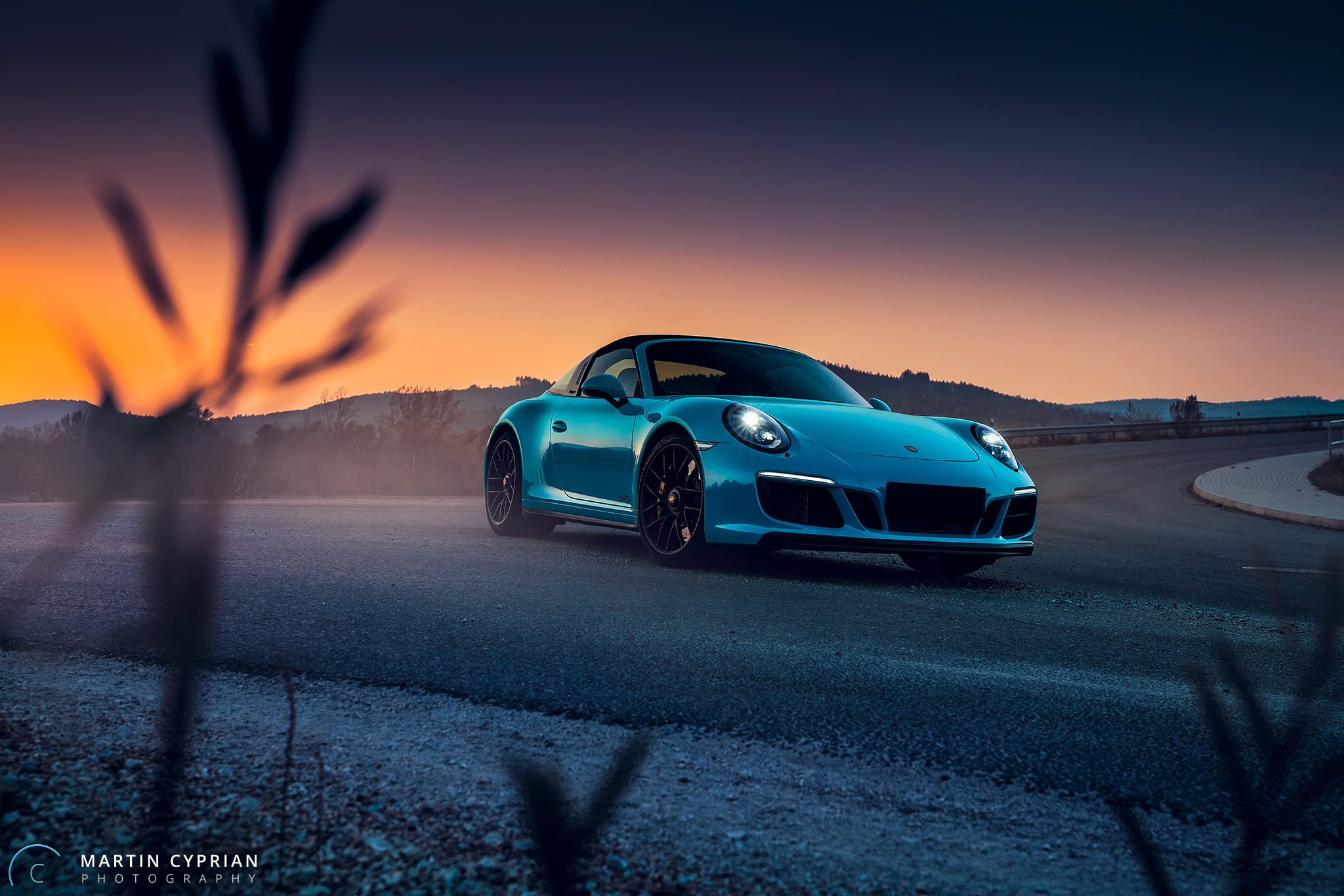 A blue Porsche 911 drives down a country road in the evening