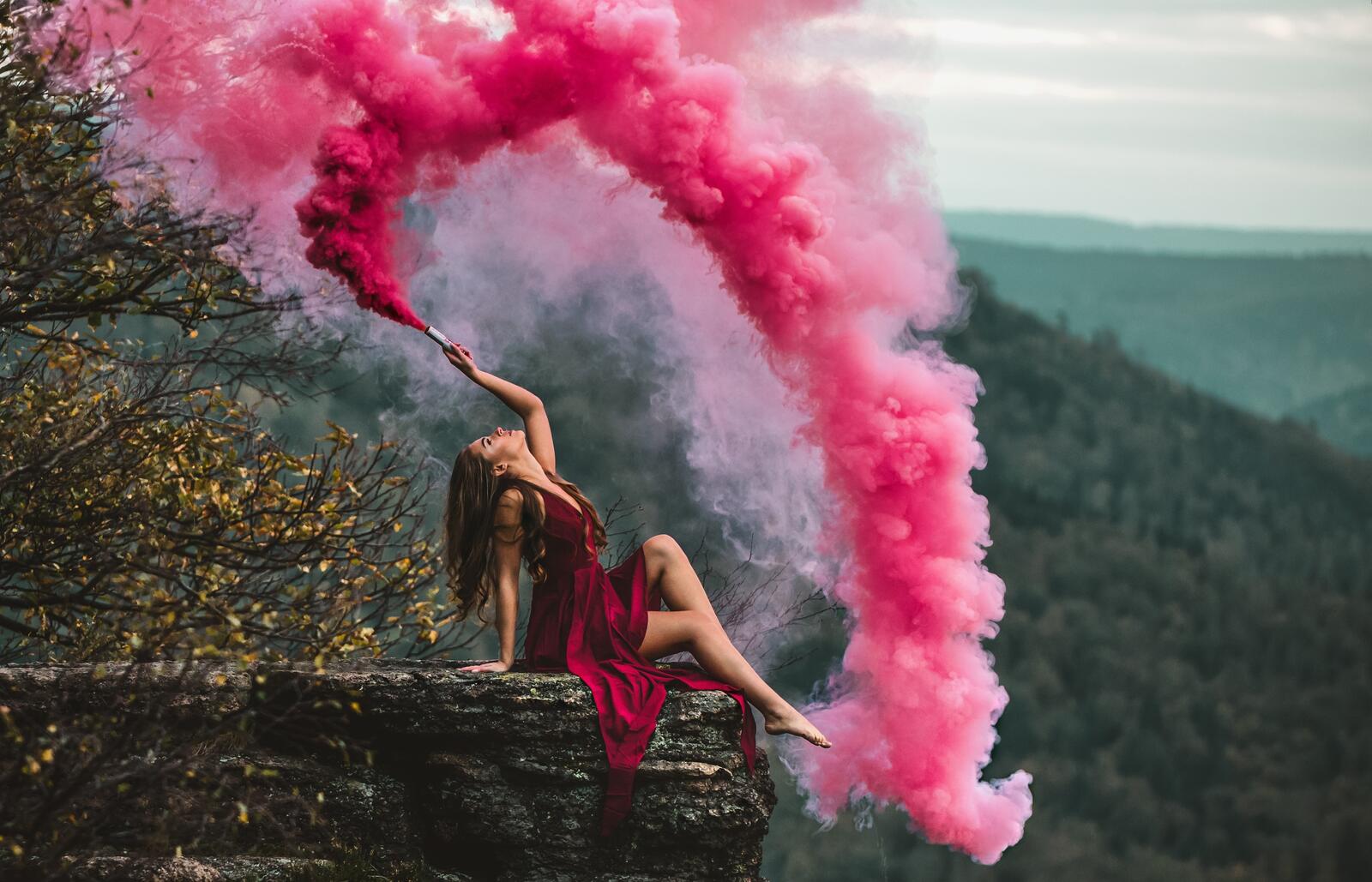 Free photo Girl sitting in a red dress on a cliff with a smoke bomb