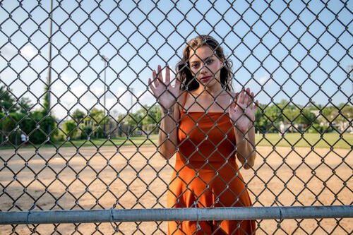 Девушка в оранжевом платье стоит за забором
