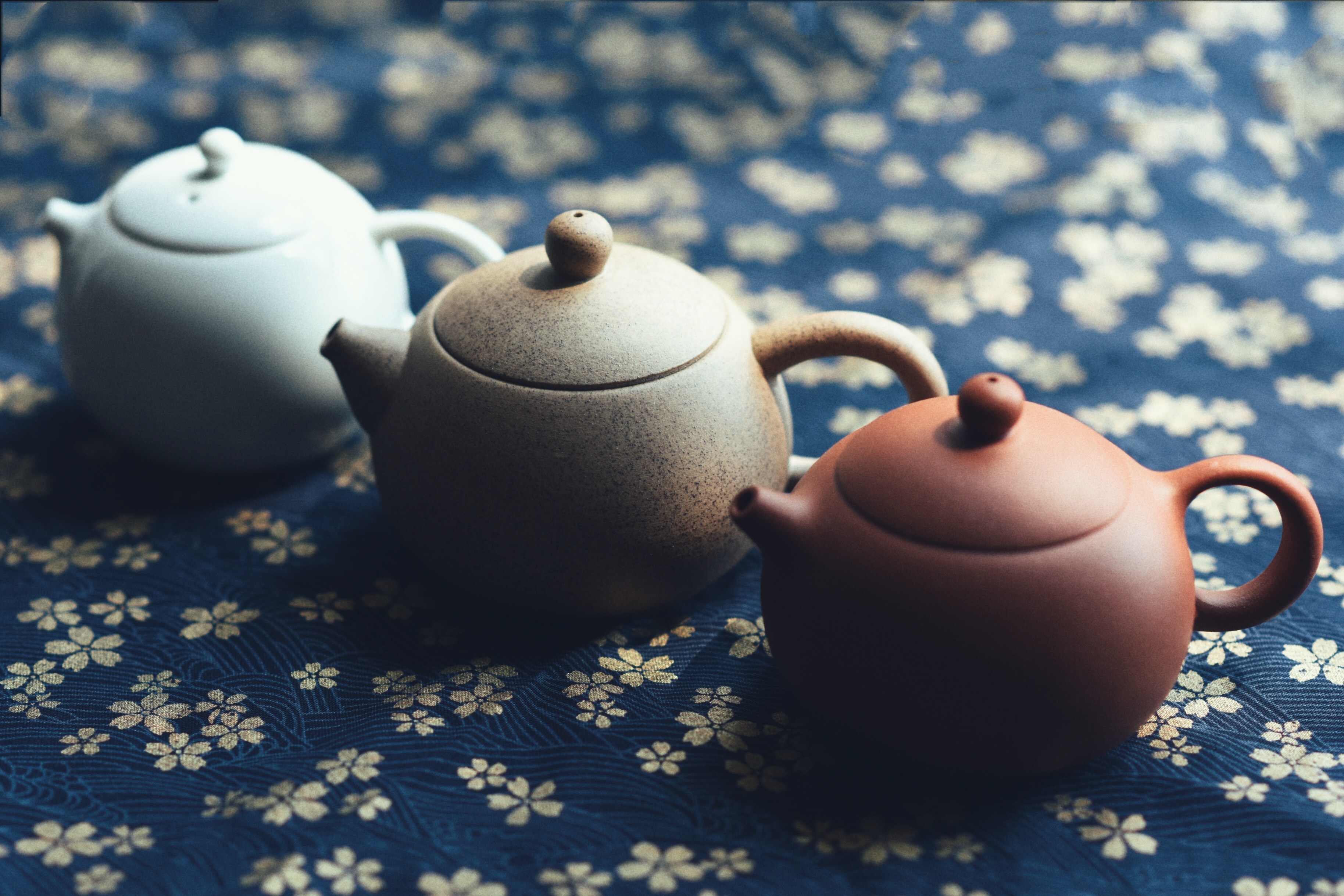 Three ceramic teapots stand on a blue fabric with florals