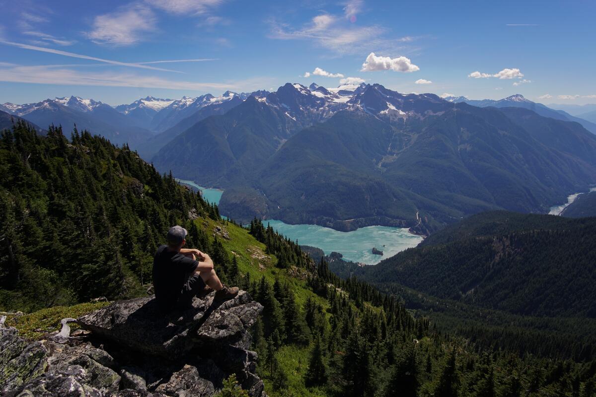 A man sits on the edge of a cliff and looks at the mountains in the distance