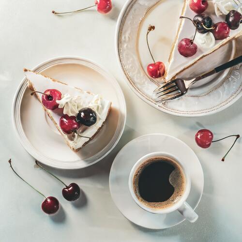 A cup of cappuccino coffee next to a plate with a slice of cream cake.