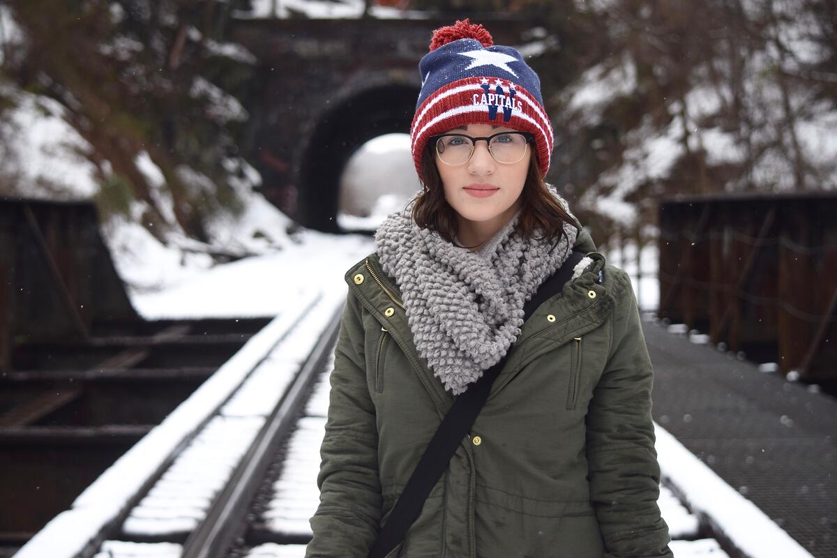 Девушка в очках и вязаной шапке зимой стоит на фоне арки в мосту