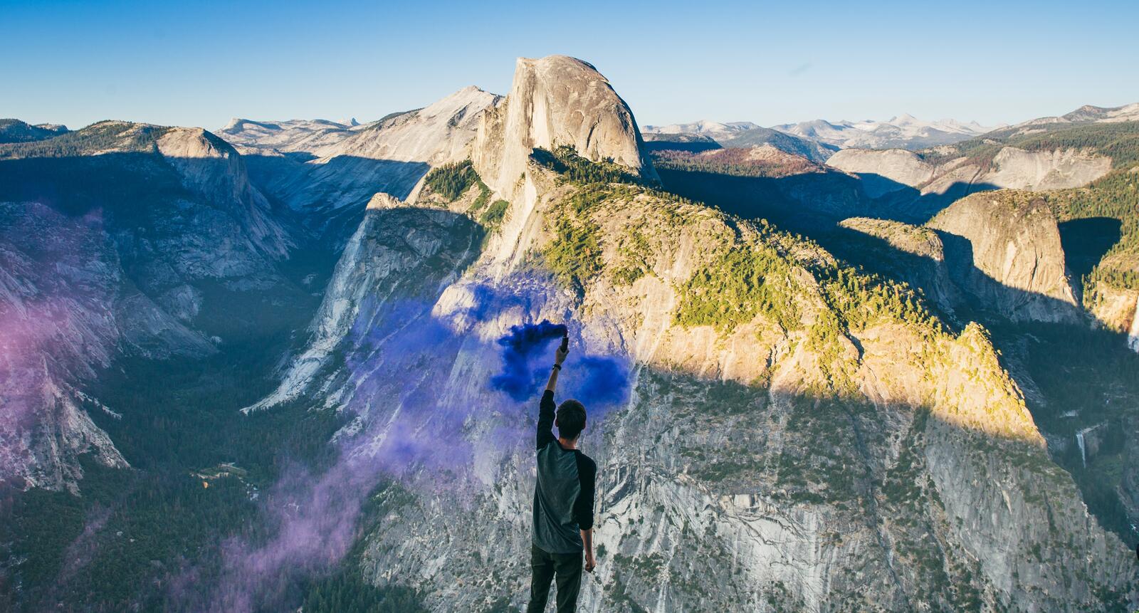 Free photo A young guy is standing on a cliff with a blue smoke bomb