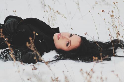 Черноволосая девушка в черном пальто лежит на снегу