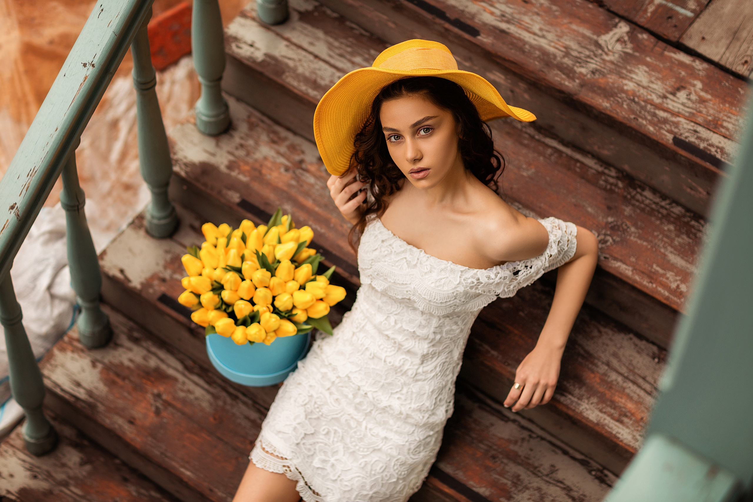 Free photo A girl in a white dress with a yellow hat on her head sits on the steps with a bouquet of yellow tulips