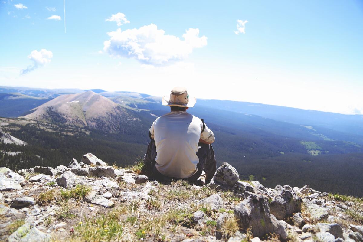 A lone man in a hat sits on a cliff edge and stares off into the distance