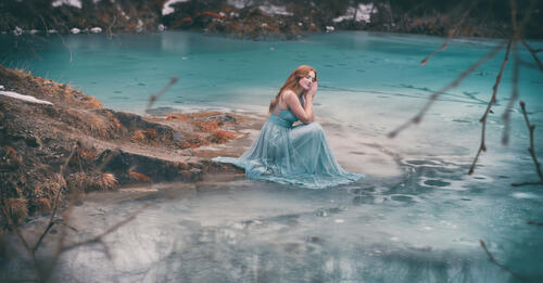 Рыжеволосая девушка в голубом платье сидит у голубого озера
