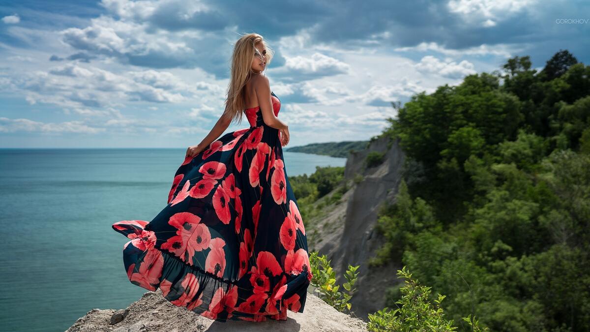 Светловолосая девушка в платье стоит на краю скалы