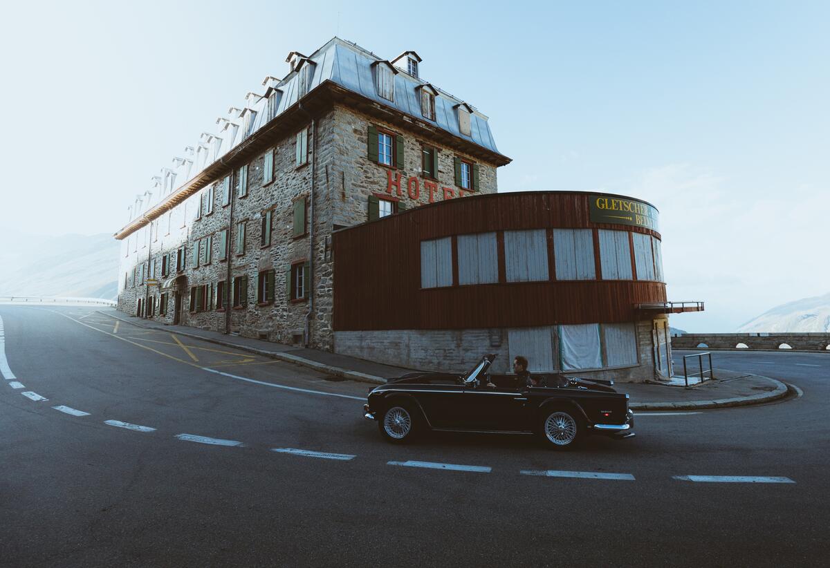 A vintage convertible drives down a city road near a building