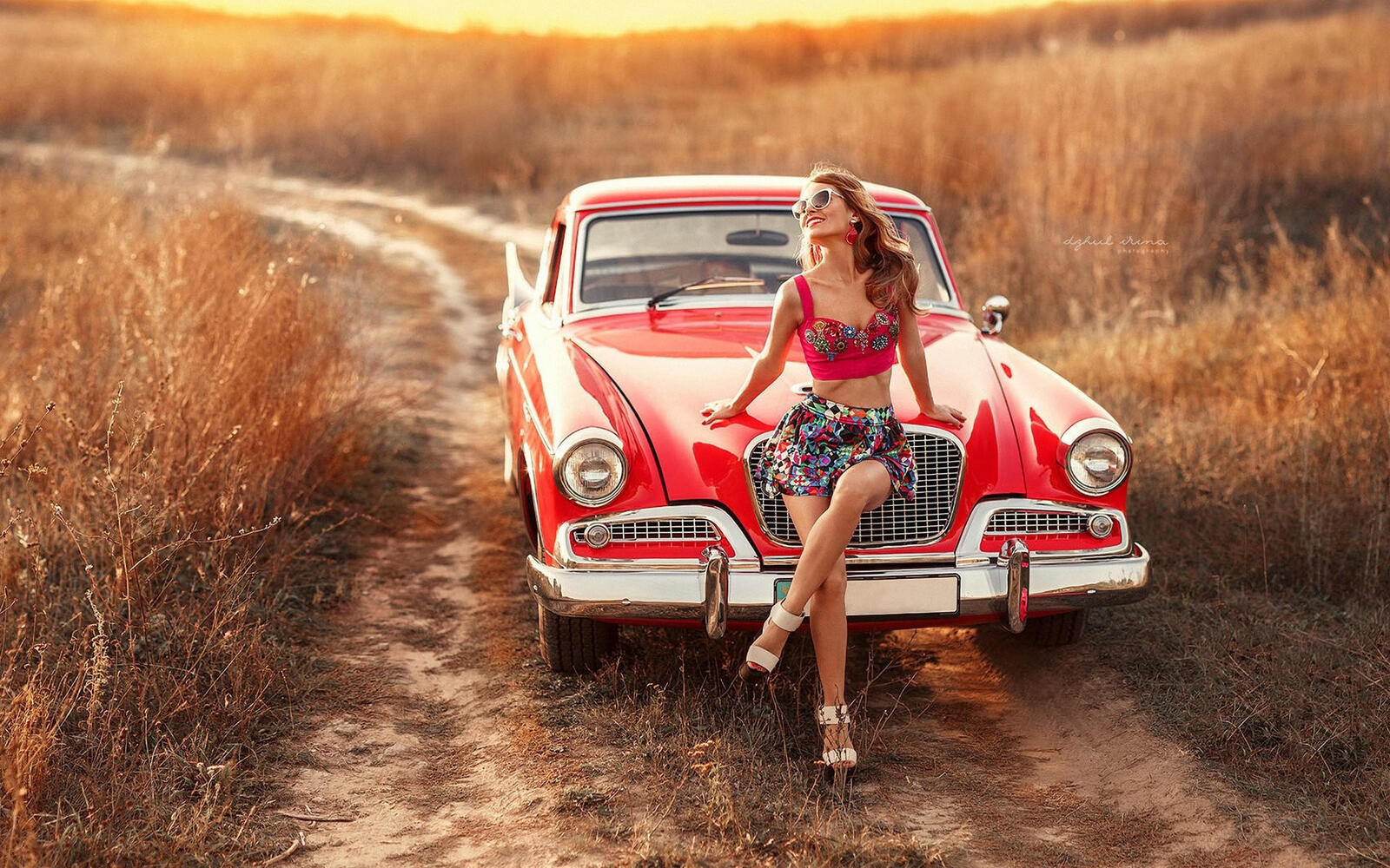 Бесплатное фото Молодая девушка в солнечных очках сидит на капоте красного автомобиля