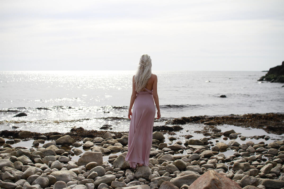 A blond girl in a pink dress stands with her back to the sea horizon