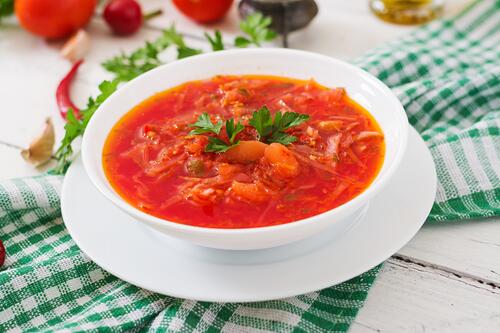 Красный суп с овощами в белой тарелке с блюдцем
