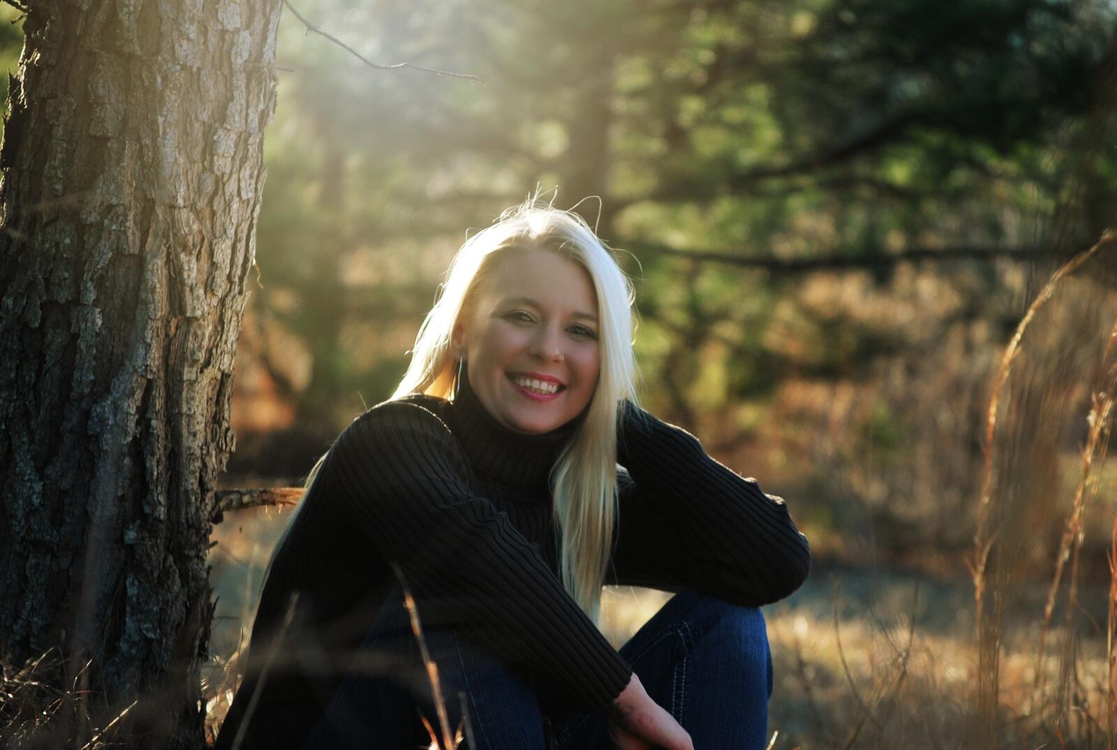 Бесплатное фото Крашенная блондинка в черном свитере улыбается сидя у дерева