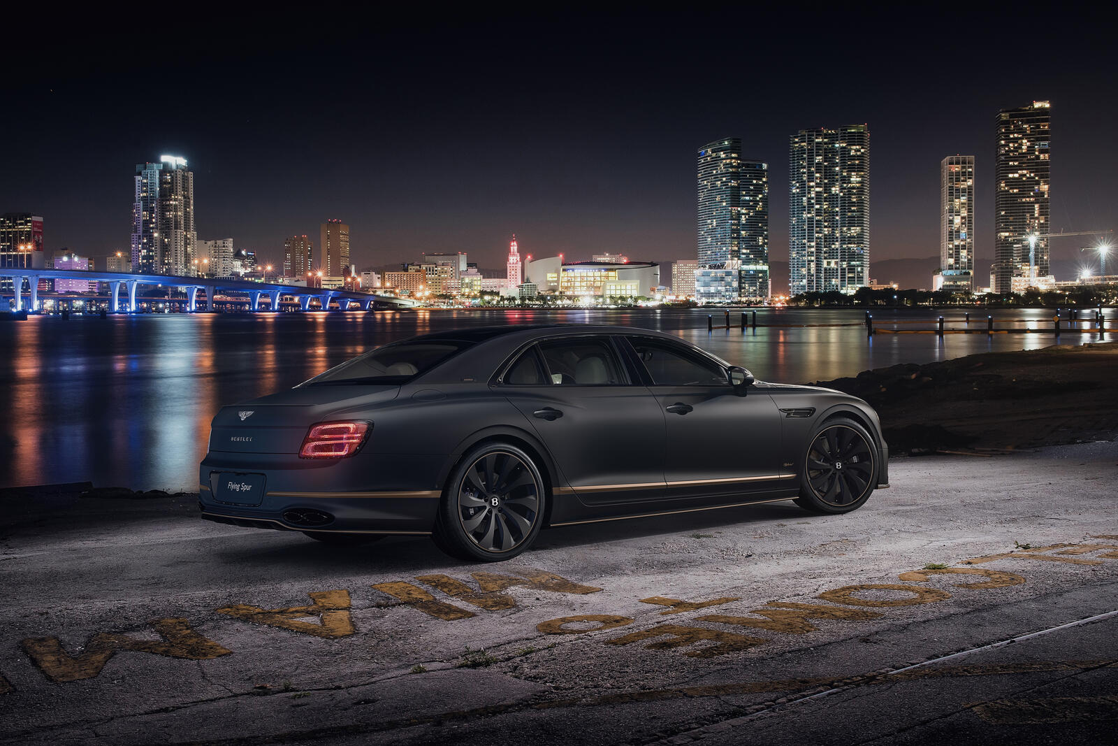 Free photo Black matte Bentley Flying Spur with gold inlays stands by the water against the backdrop of the city at night
