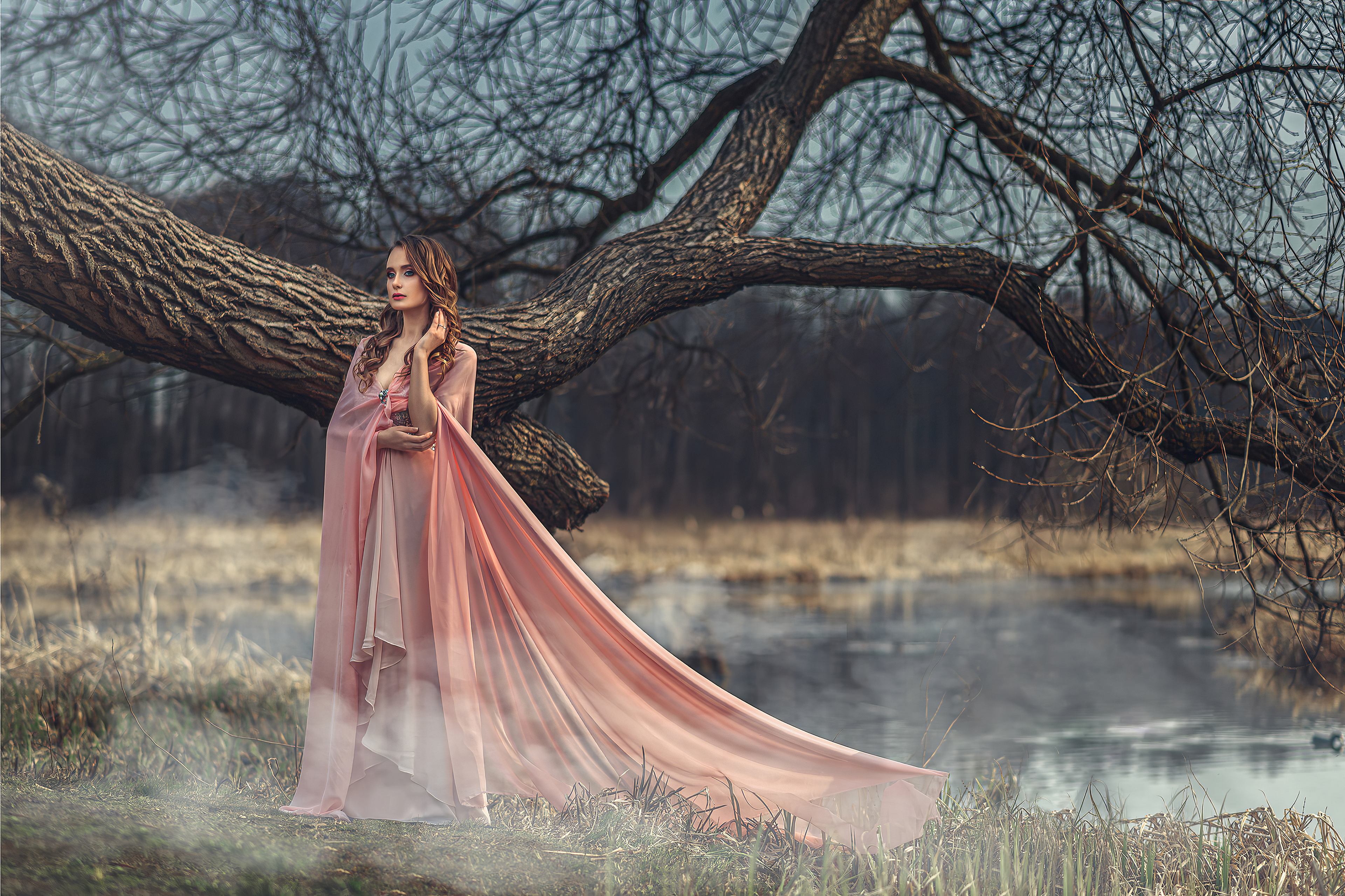 Красивая брюнетка с завитыми темными волосами стоит у ветки дерева в розовом платье на берегу озеро